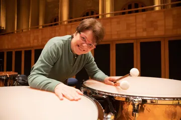 Foto de Elizabeth del Grand, mulher branca e adulta. Ela tem cabelos curtos e castanho, usa óculos retangulares e vermelhos. Ela está segurando duas baquetas e está sorrindo debruçada sobre dois tímpanos.