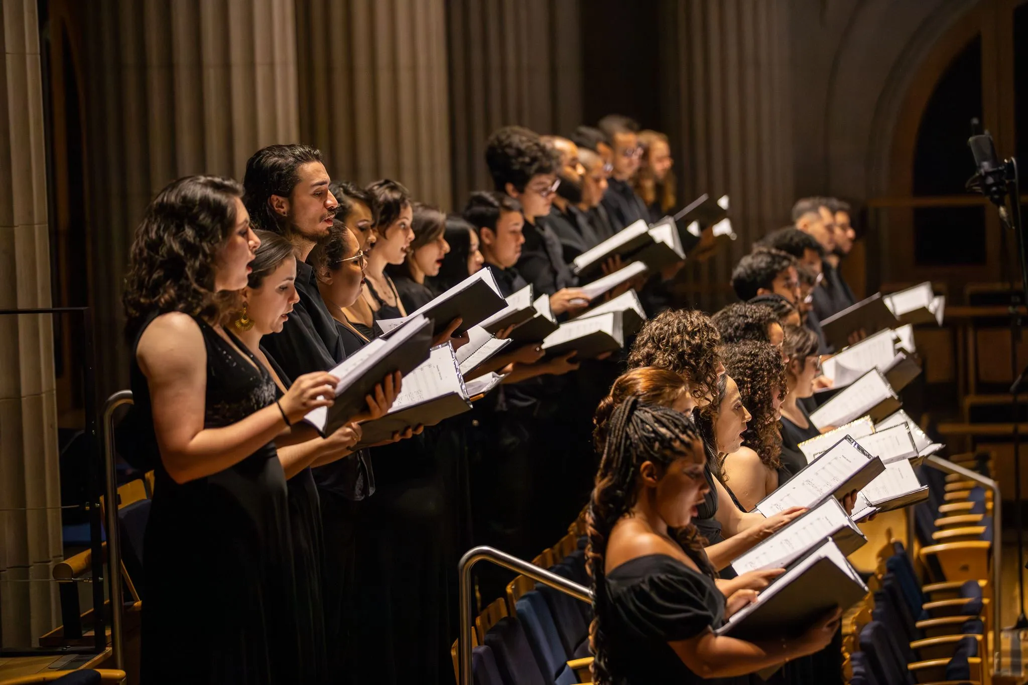 The musicians of the choir are positioned on stage. They are all wearing black and holding sheet music.