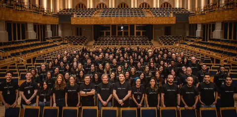 Foto dos funcionários da Fundação Osesp na sala de concertos. Eles estão de pé, dentro das fileiras de assentos. Todos vestem camisetas pretas onde se lê "osesp"