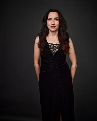 Irina Kodin is a white woman with long brown hair. She is wearing a black dress and red lipstick.