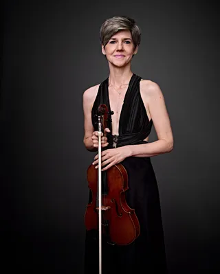 Olga Vassilevich is a white woman with short gray hair. She is wearing a sleeveless low-cut black dress and holding a viola.
