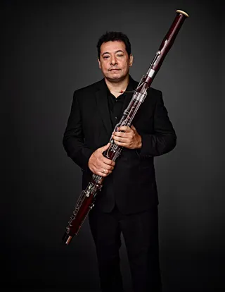 Alexandre Silvério is a white man with black hair and eyes. He is wearing a black shirt and blazer and holding a bassoon.