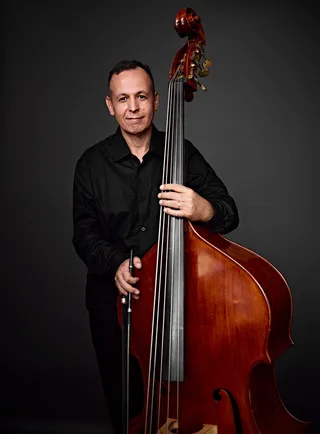 Almir Amarante é um homem branco, de cabelos e olhos castanhos. Ele veste uma camisa preta e segura um contrabaixo.