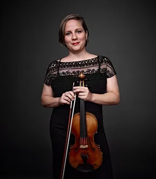 Amanda Martins is a white woman with short blonde hair and blue eyes. She is wearing a black lace top and holding a violin.