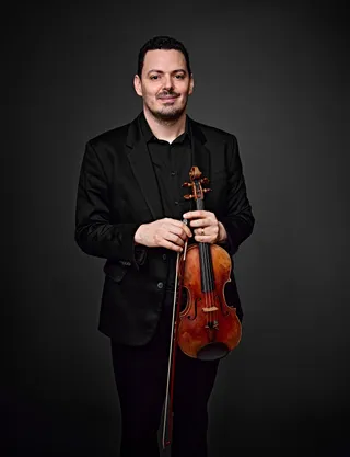 Anderson Farinelli é um homem branco, de cabelos e barba pretos. Ele veste camisa e paletó pretos e segura um violino.