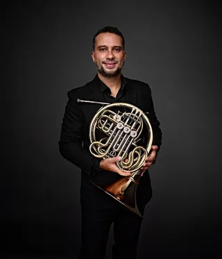 André Gonçalves is a white man with a beard and black hair. He is wearing a black shirt and holding a horn.