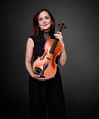 Maria Angelica Cameron is a white woman with medium red hair. She is wearing a black high-neck dress with transparency and holding a viola.