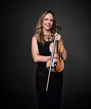 Carolina Kliemann is a white woman with long blonde hair. She is wearing a black dress with embellishments and holding a violin.