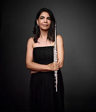 Claudia Nascimento is a white woman with brown hair and brown eyes. She is wearing a black dress and holding a flute.