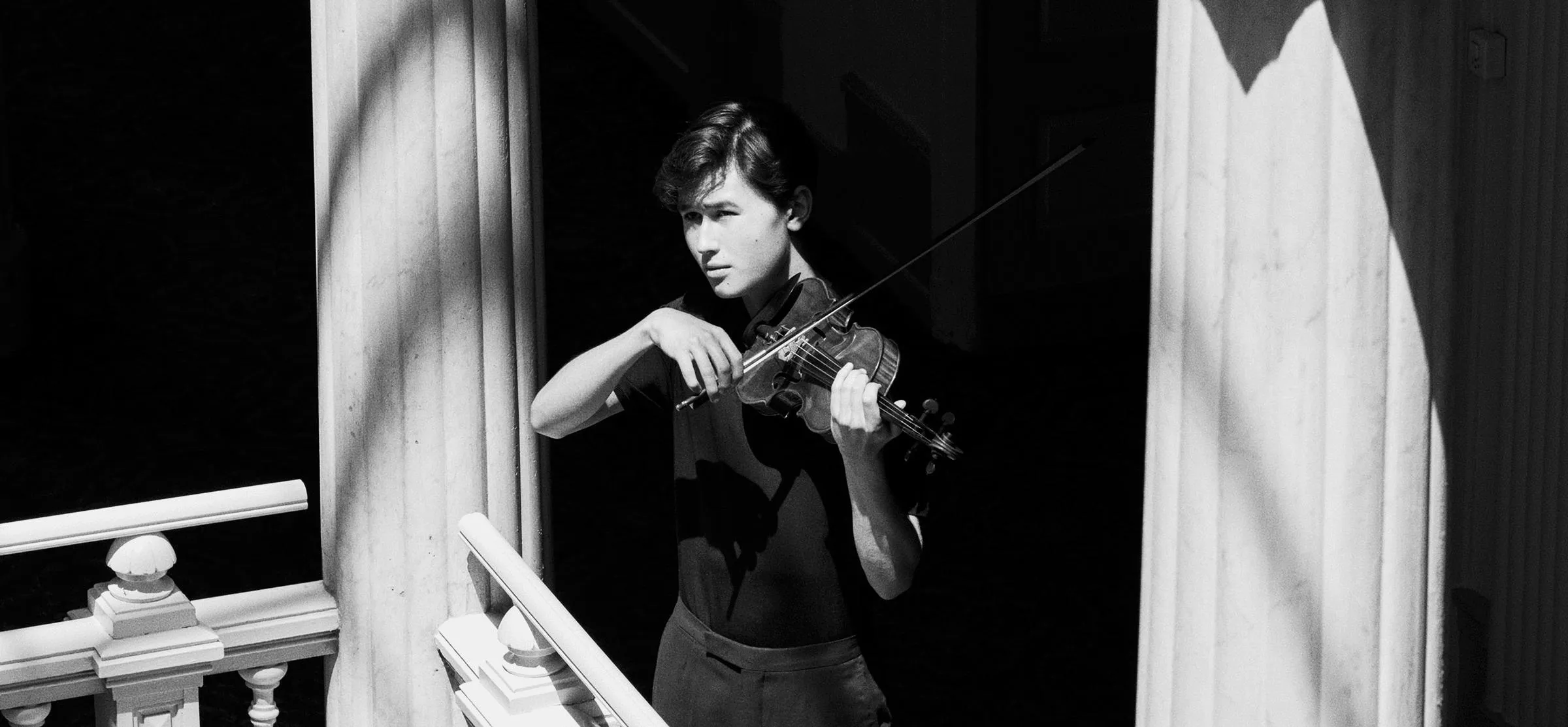 Foto em preto e branco do jovem violinista Daniel Lozakovich. Ele está segurando um violino entre duas colunas com o sol batendo em seu rosto.