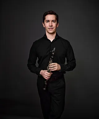 Daniel Rosas is a white man with black hair and eyes. He is wearing a black shirt and holding a clarinet.