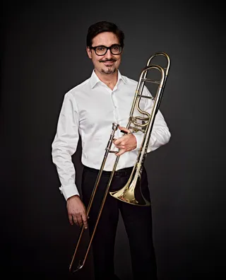 Darcio Gianelli é um homem branco, com bigode e cabelo castanho curto. Ele usa óculos, está vestido com uma camisa branca e segurando seu trombone.
