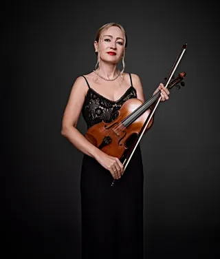 Elena Klementieva is a white woman with her blonde hair tied up and wearing red lipstick. She is wearing a spaghetti strap dress, a pearl necklace, and holding a violin bow.