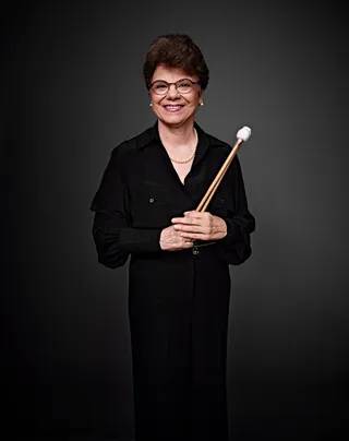 Elizabeth Del Grande is a white woman with short hair and glasses. She is wearing a black shirt and holding drumsticks.