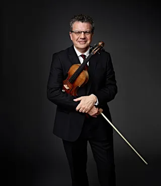 Emmanuele Baldini is a white man with glasses and gray hair. He is wearing a suit, a red tie, and a white shirt, and he is holding a violin.