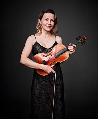 Galina Rakhimova is a white woman with shoulder-length blonde hair. She is wearing a lace dress with straps and holding a viola.