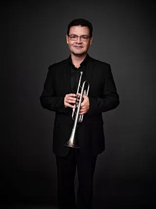 Marcelo Matos is a white man with black hair and glasses. He is wearing a black shirt and blazer and holding a trumpet.