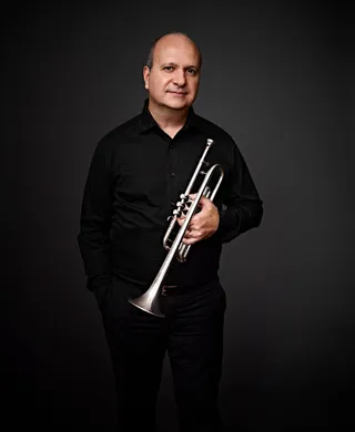 Marcos Motta é um homem branco e calvo. Ele está vestido com uma camisa preta e segurando um trompete.