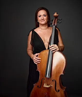 Marialbi Trisolio é uma mulher branca, de cabelos castanhos longos. Ela está usando um vestido preto decotado e segurando um violoncelo.