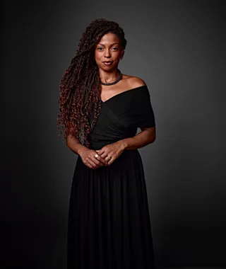 Marina Moura é uma mulher negra, com cabelo preto, longo e cacheado. Ela está usando um vestido preto com o ombro esquerdo parcialmente à mostra e um colar preto ao redor do pescoço.