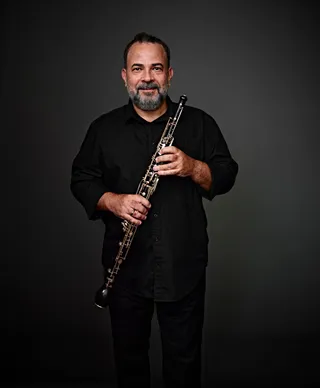 Natan Albuquerque is a white man with black hair and a gray beard. He is wearing a black shirt and holding an oboe.