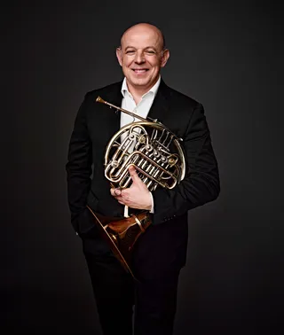 Nikolay Genov é um homem branco, calvo. Ele sorri, veste uma camisa branca e paletó preto, e segura uma trompa.