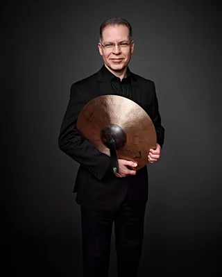 Ricardo Righini is a white man with gray hair. He is wearing glasses, a black shirt, and holding a cymbal.
