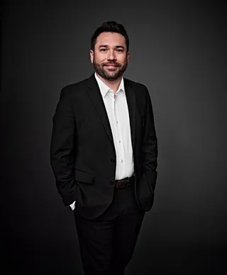 Rubén Zúñiga is a white man with brown hair and a beard. He is wearing a white shirt and a black blazer, and he is holding drumsticks.