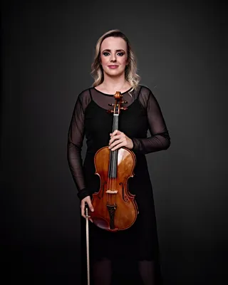 Sarah Pires é uma mulher branca, de cabelos loiros e longos. Ela está usando um vestido preto de mangas compridas com transparência e segurando uma viola.