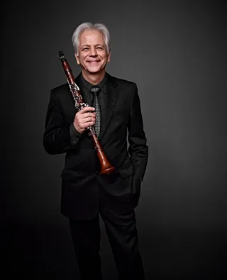 Sergio Burgani é um homem branco, com cabelo grisalho. Ele está usando uma camisa preta e uma gravata com detalhes prateados, um blazer preto e segura um clarinete.