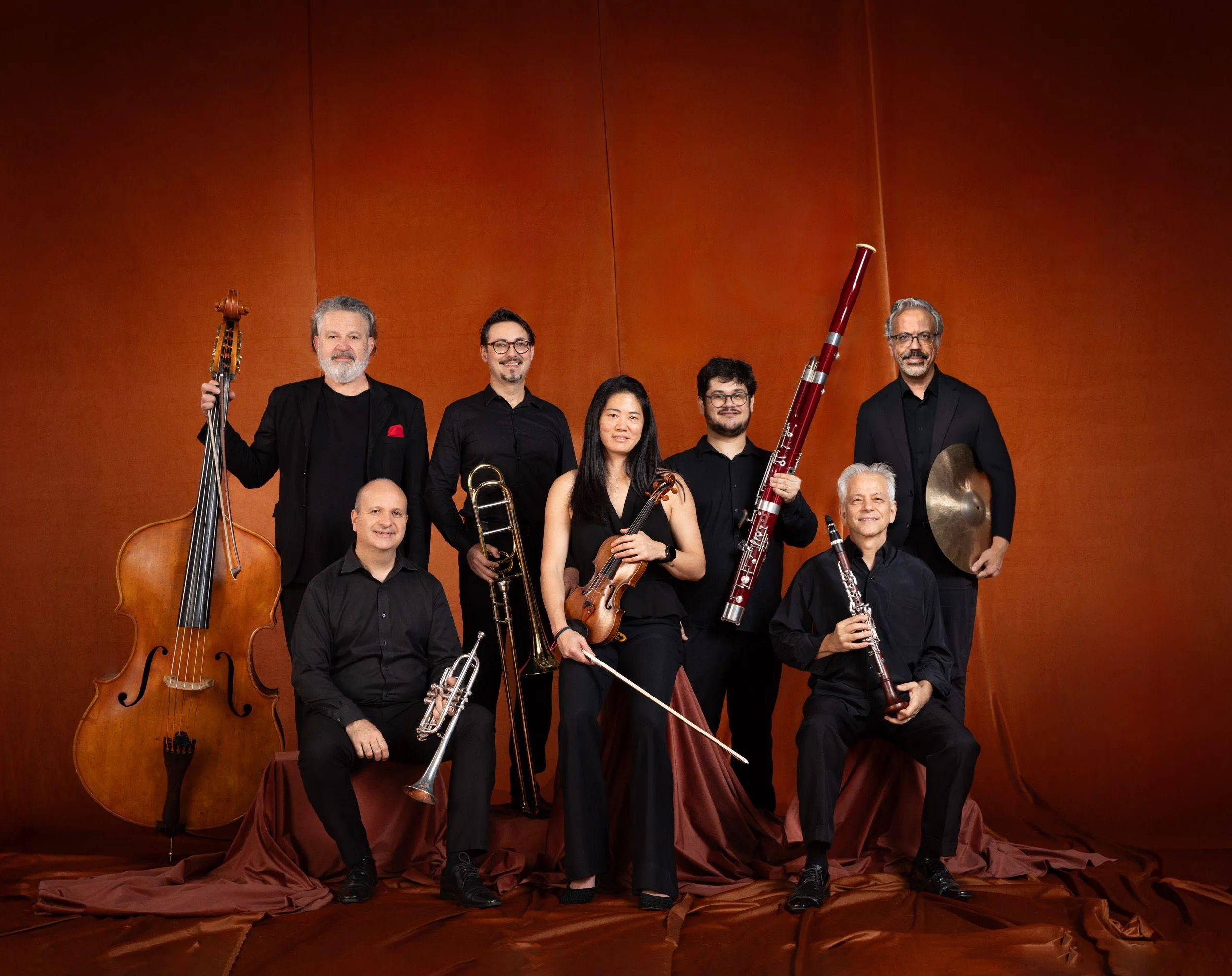 Os músicos estão sentados com seus instrumentos em frente de um tecido laranja.