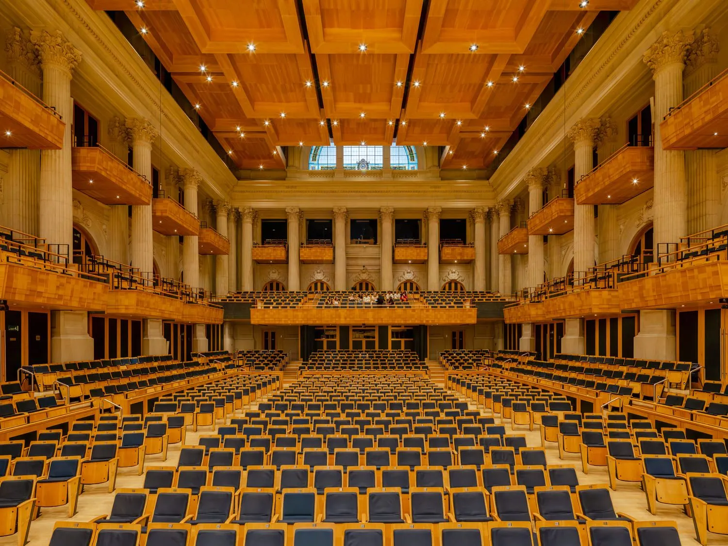 Sala de concertos com pessoas na plateia e no palco. As colunas estão iluminadas de azul e o palco com uma luz quente.