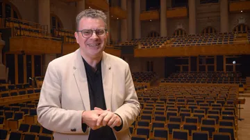 Foto de em plano médio de Emmanuele Baldini, homem branco, adulto, com cabelos grisalhos curtos e encaracolados. Veste camisa preta e um blazer bege. Ele está de pé no palco da sala de concertos.
