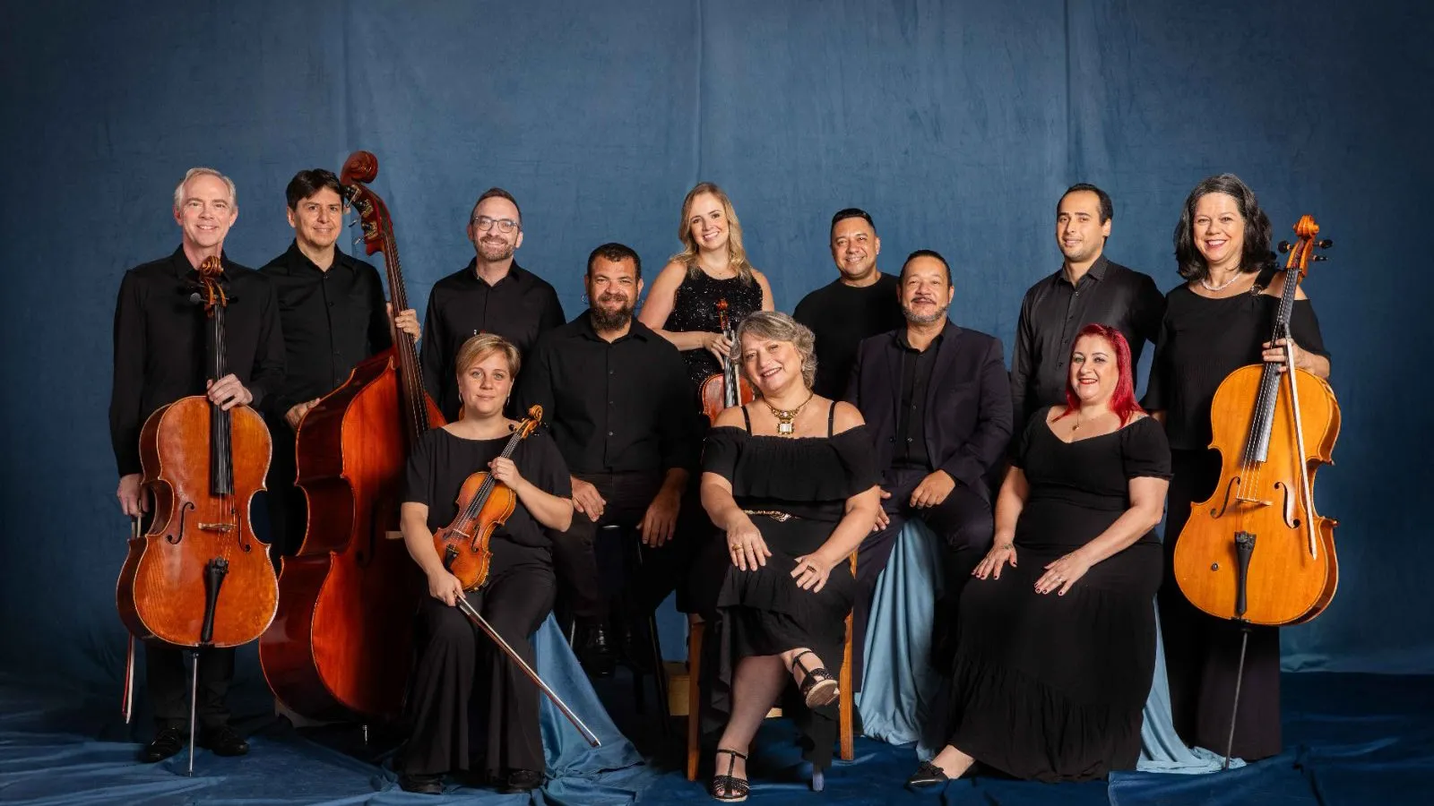 Foto de 12 músicos sentados e em pé, a frente de uma cortina azul. Todos vestem preto e olham para a foto.