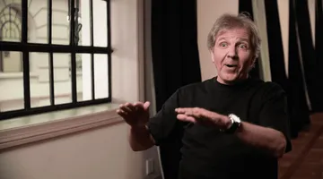 Foto do maestro Thierry Fischer. Homem branco de cabelos grisalhos e olhos claros. Veste camisa manga longa preta e um relógio prata no pulso esquerdo. Está sentado ao lado de uma janela.