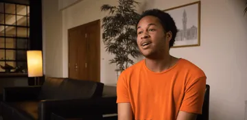 Foto de Sheku Kanneh-Mason, homem preto e de cabelos crespos e curtos. Ele está sentado e veste uma camiseta laranja bem vibrante.