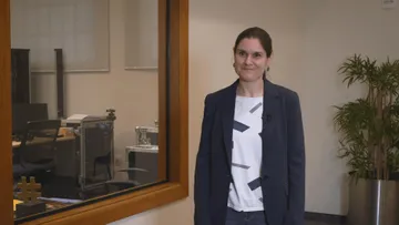 Delyana Lazarova está em pé em um ambiente interno, sorrindo. Ela veste blazer azul-marinho, camiseta branca estampada e calça jeans. Ao fundo, há uma janela de vidro que revela uma sala com equipamentos e, à direita, uma planta em vaso metálico.