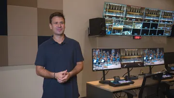 Foto de Cristian Budu, um homem branco com cabelo castanho curto. Ele está no estúdio audiovisual da Sala São Paulo, é possível ver atrás dele diversas telas dedicadas à transmissão de um concerto.