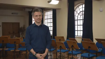 Foto de Joseph Bastian, homem branco de cabelos grisalhos. Ele veste uma camisa azul e esta segurando as mãoes em frente ao corpo.