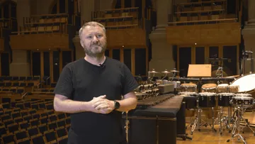 Colin é um branco de barba curta e cabelo claro, vestindo camiseta preta, está em pé no palco de uma sala de concertos vazia. Atrás dele, há uma grande variedade de instrumentos de percussão organizados, incluindo tambores e xilofones.