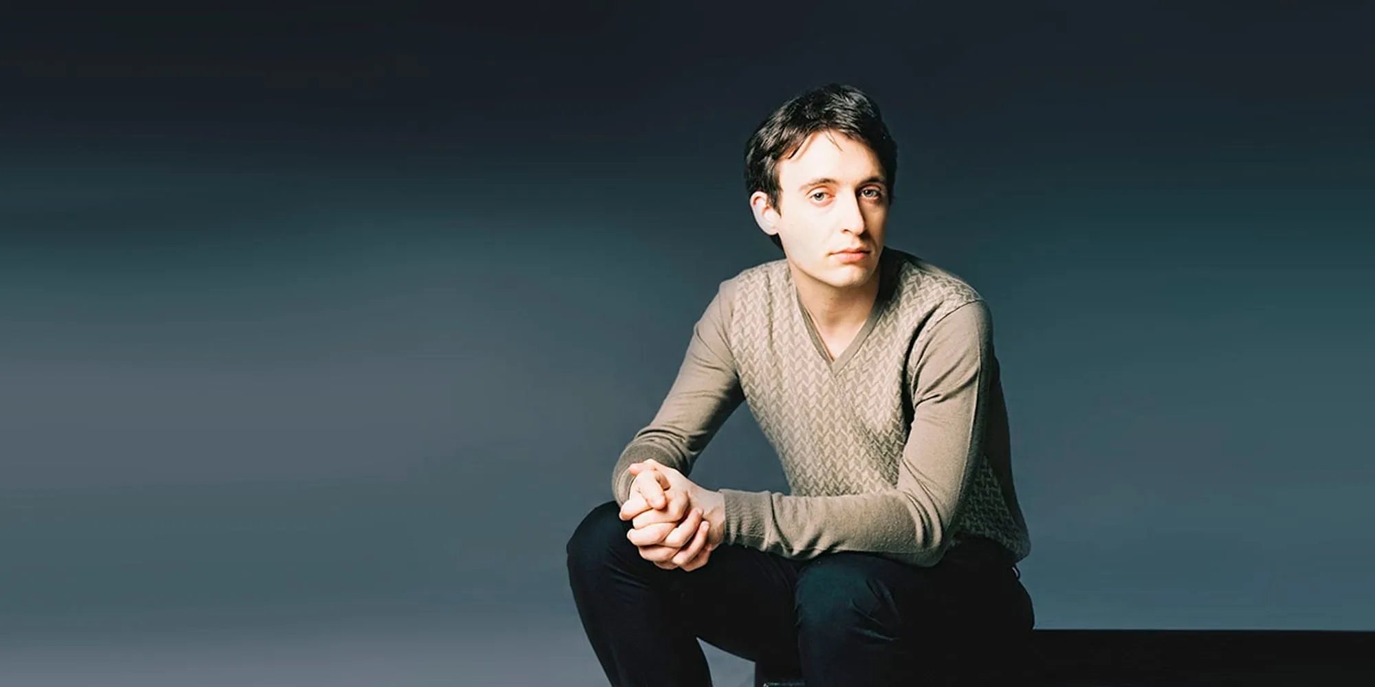 Foto de Jean-Frédéric Neuburger, homem branco e adulto. Ele possui cabelos castanhos e curtos. Está sentado sobre uma caixa preta. Ele olha para a camera e segura uma mão na outra, com ombros sobrepostos aos joelhos.
