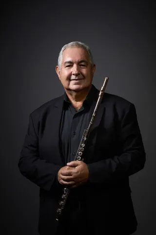 Savio Araujo is a white man with short gray hair. He is wearing a black shirt and holding the flute close to his chest.
