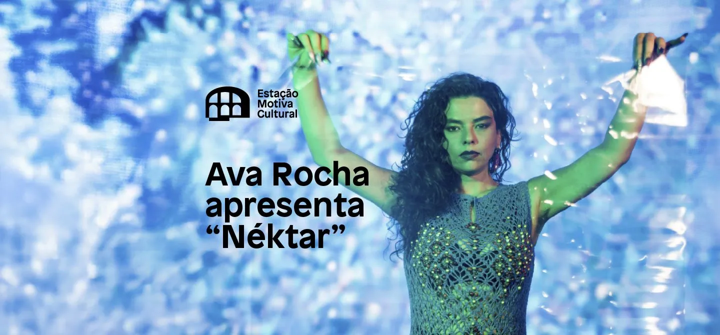 A imagem mostra Ava Rocha, uma mulher branca de cabelos longos pretos, de mãos para cima e na frente de um fundo azul.