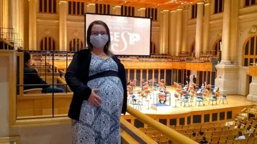 Camila é uma mulher branca, ela está grávida e posa na Sala de Concertos usando uma máscara cirúrgica.